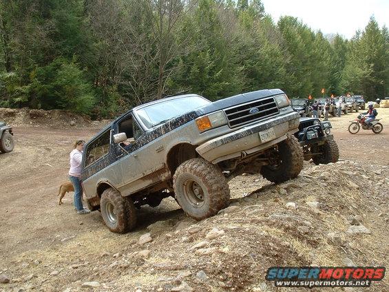 Mud-splattered Ford Bronco climbing a dirt hill, front 3/4 view
