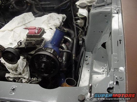 Engine bay with blue turbocharger intake pipe, red ignition coil, and pulleys. Some white cloths cover parts of the engine