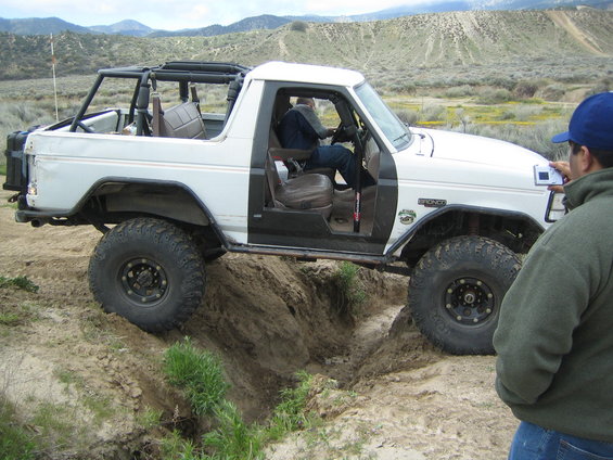 White Ford Bronco with roll cage driving over dirt obstacle. Person filming with camera