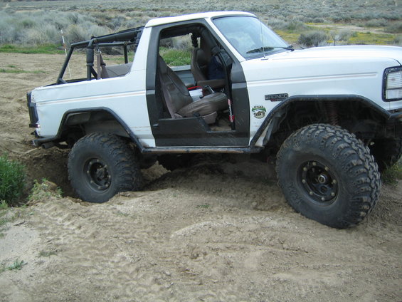 White 1987 Ford Bronco with off-road tires stuck in sand, driver's door open
