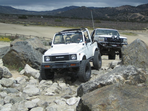 White Suzuki Samurai and a white truck driving over rocks. The Samurai has 'The Web Wheelerz' on its roof rack