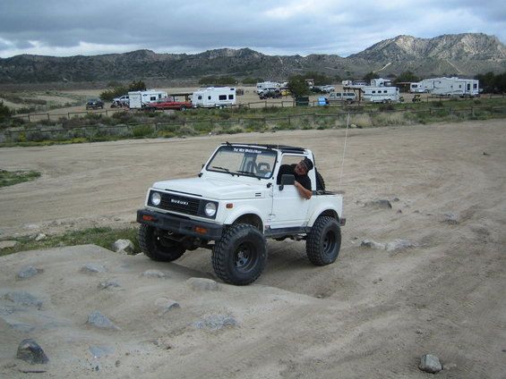 1987 Suzuki Samurai with lifted suspension and large tires on a dirt trail. Driver is leaning out the window
