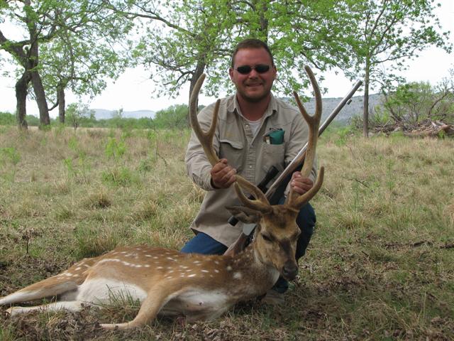 Man kneeling in grassy field holding deer antlers, rifle nearby