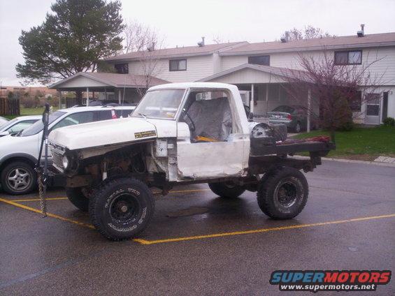 White pickup truck with flatbed, missing doors and fenders, parked in a lot