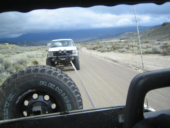 White pickup truck with aftermarket bumper and lights being towed on a dirt road. Spare tire visible in foreground