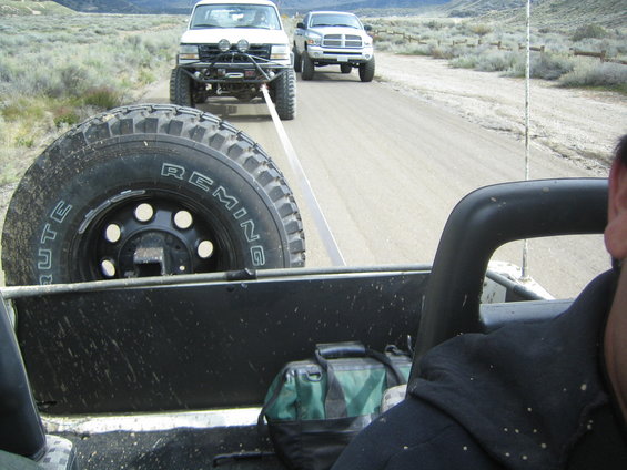 White off-road truck being towed by a second vehicle, with a Dodge Ram following behind on a dirt road