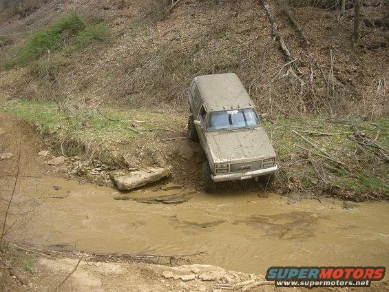 Muddy SUV driving through a creek bed on a wooded trail. The vehicle appears to be a GMC Jimmy from the late 1980s