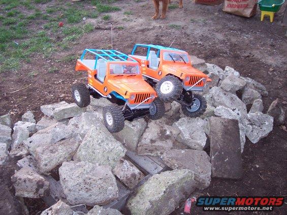 Two orange remote-controlled jeeps with blue roll cages are climbing over a pile of rocks