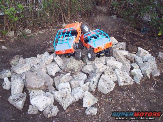 Two orange toy trucks with blue roll cages are piled on rocks