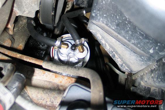 Fuel filter with brass fittings and black hoses attached, surrounded by engine components and rust