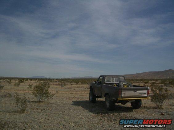 Pickup truck parked in a dry, desert landscape with sparse bushes and mountains in the distance. Spare tire is visible in the truck bed