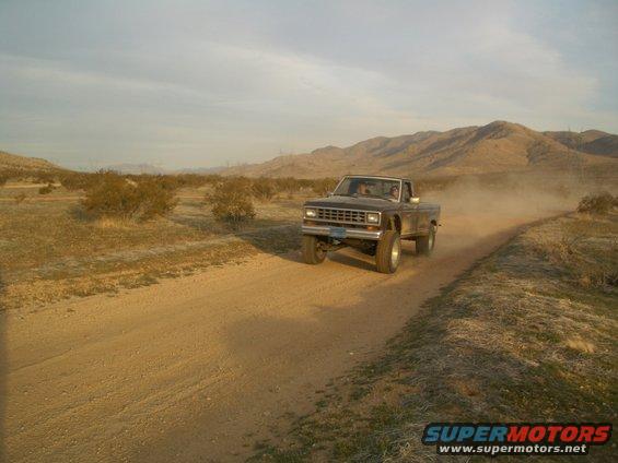1985 Ford Ranger pickup truck driving on a dusty dirt road in a desert landscape