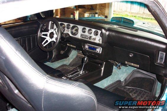 Car interior with black leather seats, dashboard with gauges, and aftermarket steering wheel. Teal exterior visible through windshield