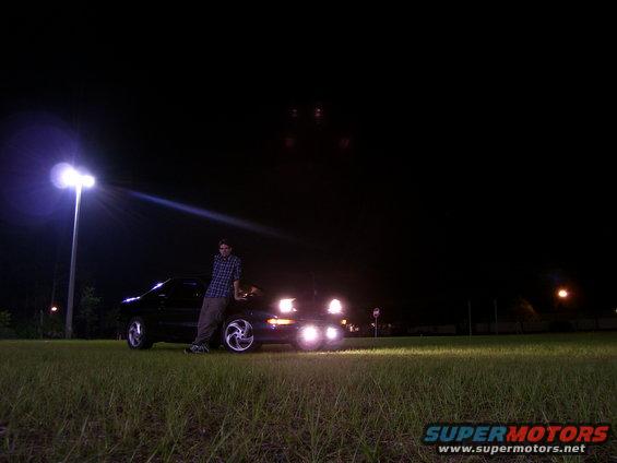 Man leaning on a black 1994 Ford Probe at night, headlights on