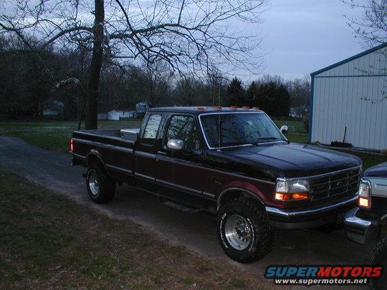 Dark red and black Ford F-Series truck, 3/4 front view, parked on a dirt road next to a white building