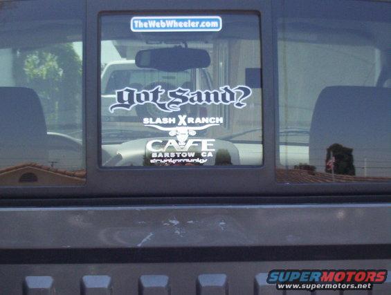 Rear window of a truck with stickers: TheWebWheeler.com, Got Sand? SLASH XRANCH, CAFE BARSTOW CA drunkemunity. Another vehicle is reflected in the window