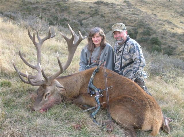 Deer with large antlers lies on the ground next to two hunters
