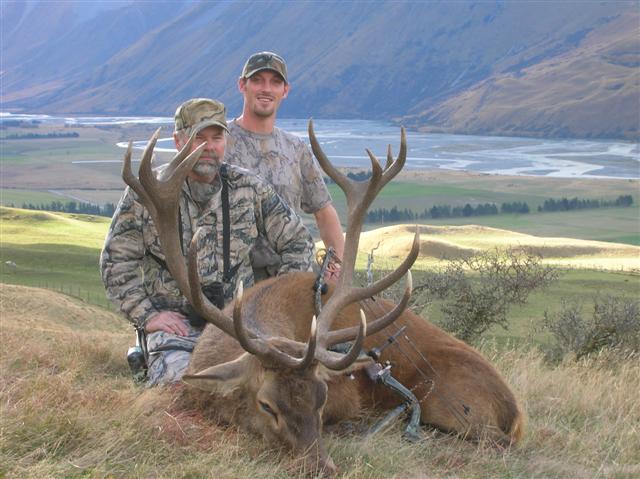 Two hunters pose with a large elk in a mountainous landscape