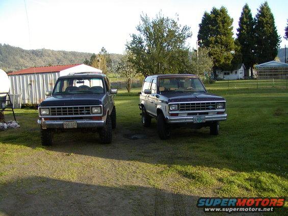 Two Ford Bronco II SUVs parked on grass, one dark blue, one two-tone blue and white