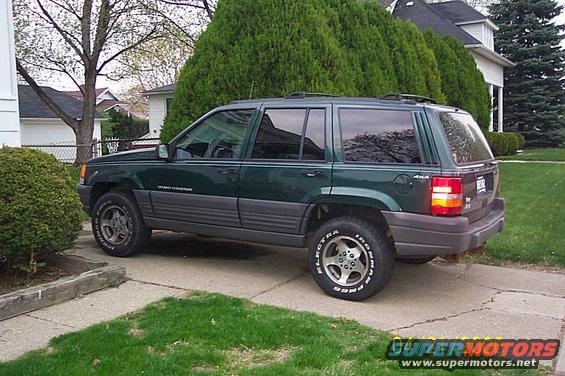 Green Jeep Grand Cherokee parked on a driveway, side view. Tires have white lettering