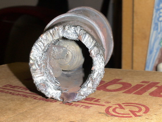 Damaged metal pipe with rough, torn edges. Cardboard box with red text is visible underneath
