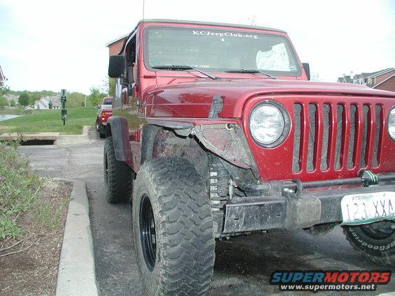Red Jeep Wrangler front quarter view, parked next to a curb. Another red vehicle is visible in the background. Sticker on windshield reads KCJeepClub.org
