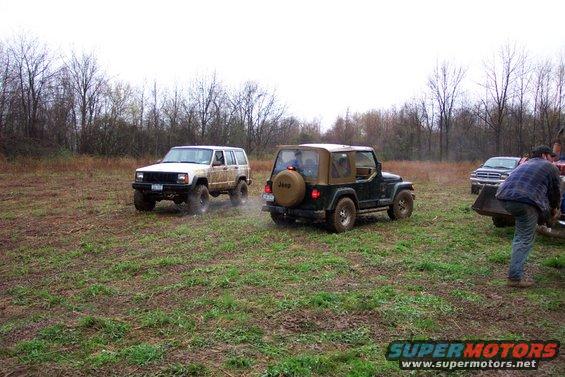 Two Jeeps driving on a muddy field with trees in the background. A person is near a truck on the right