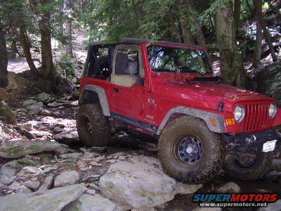 Red 2003 Jeep Wrangler navigating a rocky stream bed in a forest
