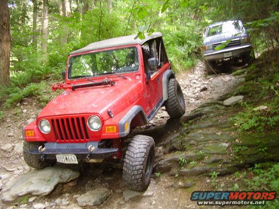 Red Jeep Wrangler navigating a rocky trail, with a dark SUV behind it on a wooded path