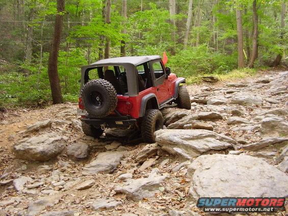 Red 2003 Jeep Wrangler with soft top, spare tire on rear, navigating a rocky forest trail