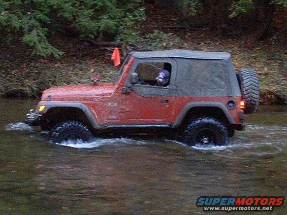 red Jeep Wrangler driving through a shallow stream, trees in background