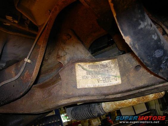 Underside of a vehicle showing rusty suspension parts and a sticker with part numbers like D-108-A and 37B-3002-RG