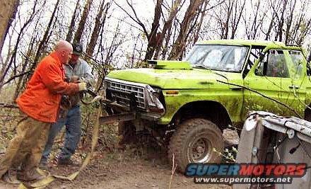 Green 1978 Ford Bronco with a winch, being pulled by two men in a wooded area