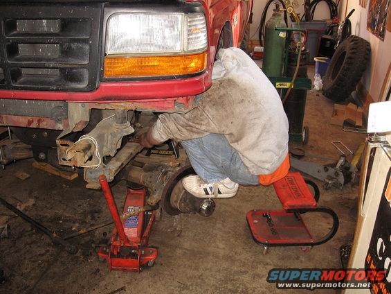 Red Ford truck front end with a person working on the suspension, jacked up in a garage