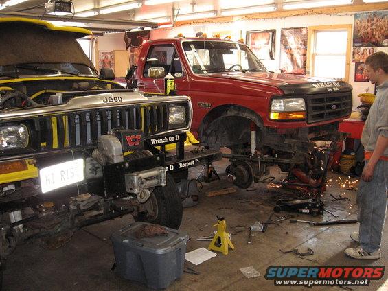 Yellow Jeep and red Ford truck in garage, both appear to be undergoing repairs or modifications