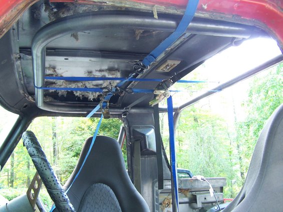 Interior view of a vehicle with a roll cage, straps, and aftermarket seats. Insulation is visible in the roof area