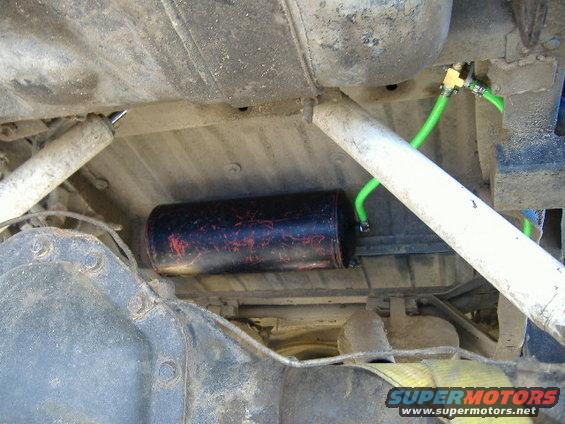 Undercarriage of a vehicle showing a black cylinder with red markings, green hoses, and suspension parts