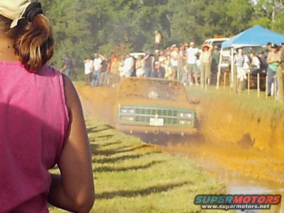 Green truck with a prominent grille driving through mud, spectators watch