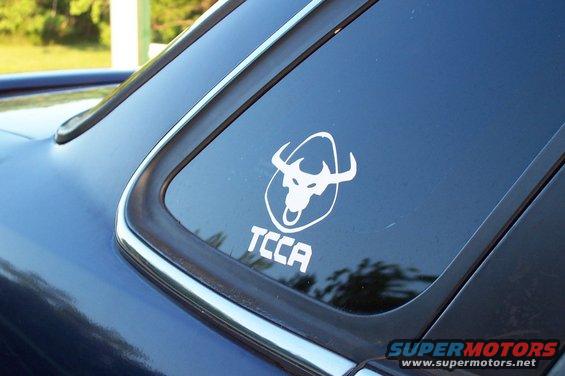 Blue car rear window with white TCCA bull logo sticker. Supermotors.net watermark at bottom right