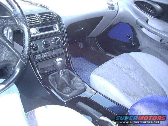 Car interior with manual shifter, dashboard, and patterned seats. "SUPERMOTORS.NET" visible