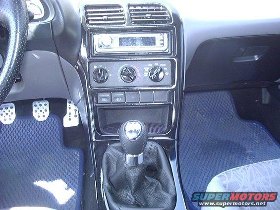 Car interior with manual gear shift, radio, and climate controls. Pedals and blue floor mats are visible