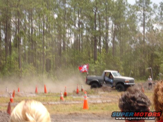 Pickup truck with a flag on the back driving on a dirt track, kicking up dust. Orange cones mark the course
