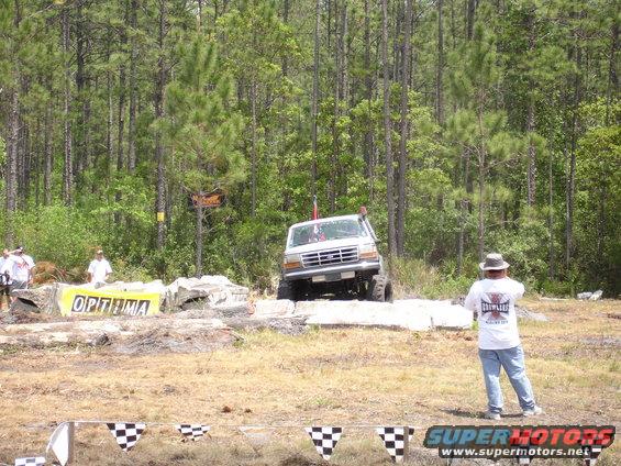 White Ford truck with lifted suspension and large tires, angled from the front-right, driving over concrete obstacles