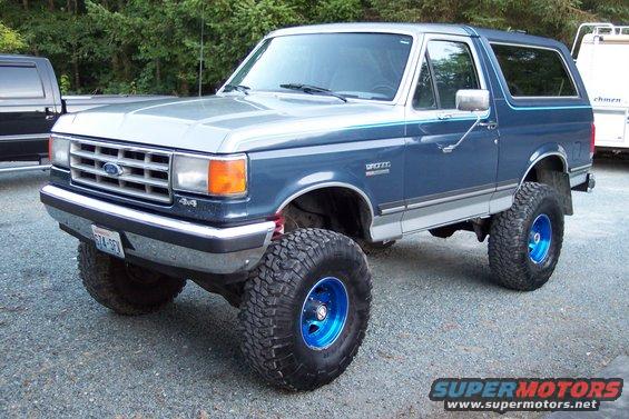 Blue and silver 1987 Ford Bronco with lifted suspension and blue rims, parked on gravel
