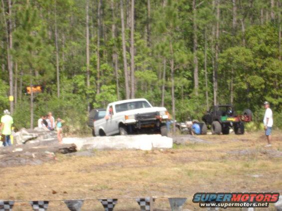 White pickup truck with a black bed liner is on a dirt obstacle course. Another black off-road vehicle is nearby