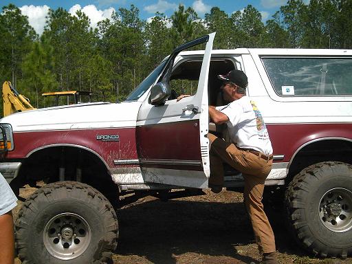 White and maroon Ford Bronco with large tires, driver's door open, man climbing in
