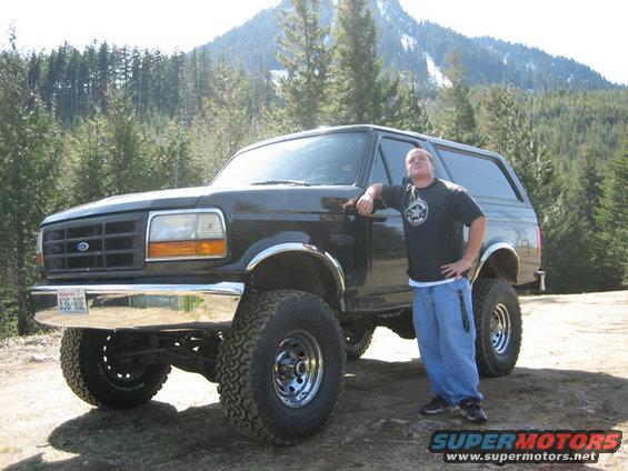 Black Ford Bronco with lifted suspension and large tires, parked on dirt with a man leaning against it. Mountains and trees in background