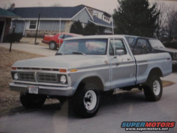1977 Ford F-150 pickup truck, silver with blue stripe, parked in driveway. Red car in background