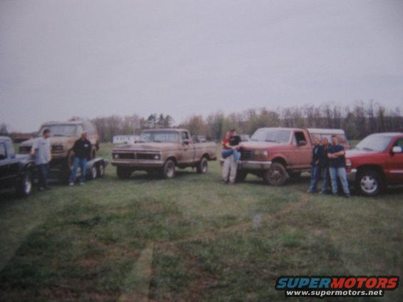 Several pickup trucks and an SUV parked in a grassy field with people standing around them