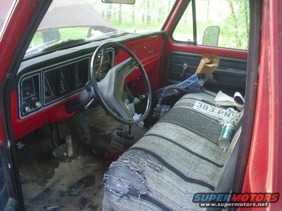 Interior of a red 1976 Ford F-100 pickup truck, showing the dashboard, steering wheel, and torn bench seat